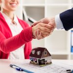 image of a woman in red sweater shaking hands on a real estate deal. There is a small model house on the table in front of her.