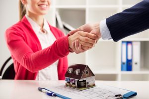 image of a woman in red sweater shaking hands on a real estate deal. There is a small model house on the table in front of her.